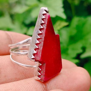 Può includere: Un anello in argento con una grande pietra rossa verticale a forma di freccia, incastonata in una montatura in metallo seghettato. La fascia è composta da tre fili d'argento sottili e paralleli collegati alla base dell'incastonatura, creando un aspetto a strati.