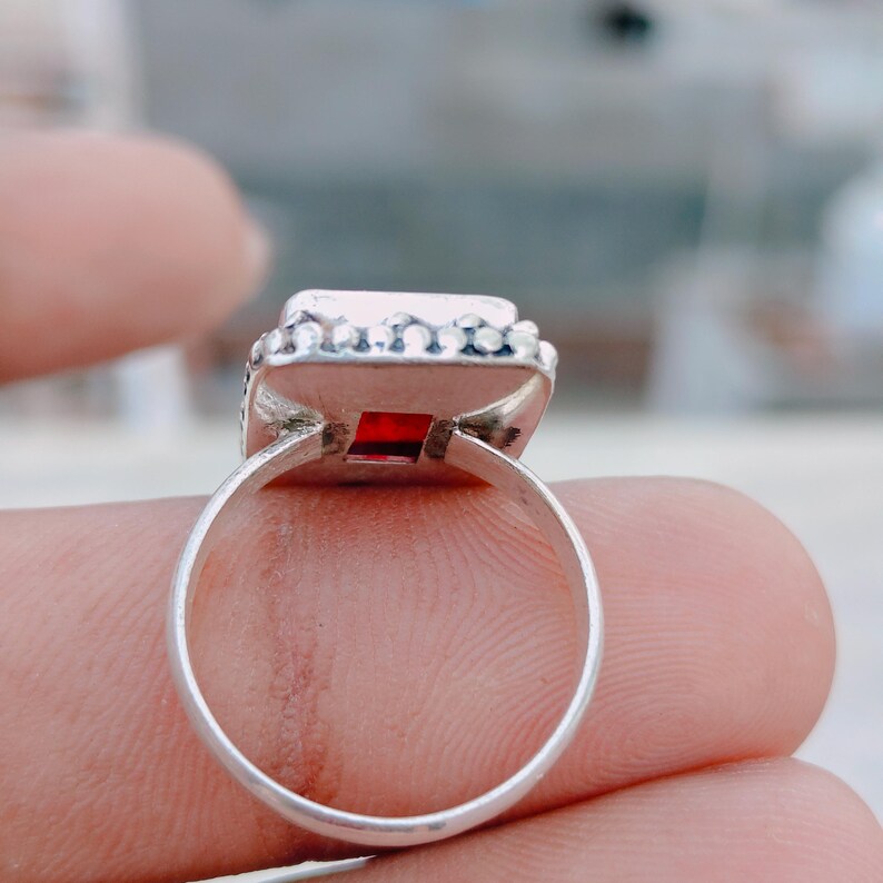 May include: A silver ring featuring a square-cut red gemstone set in a bezel. The metal band has a smooth, polished finish, and the setting is decorated with a series of small, raised silver spheres along the outer edge of the square frame.