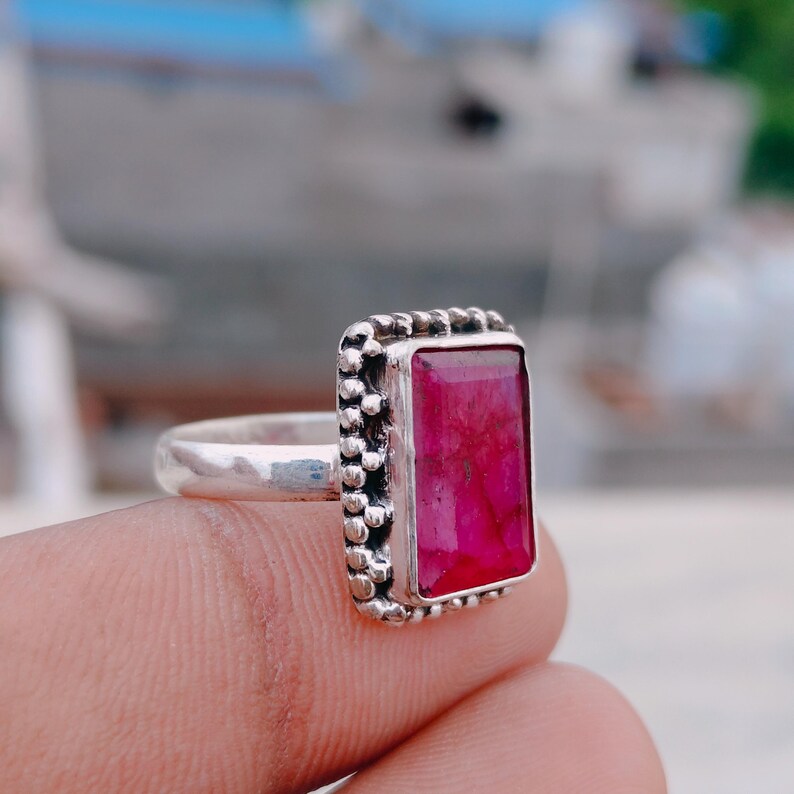 May include: A silver ring featuring a rectangular, faceted, deep pink gemstone set in a bezel. The metal setting surrounding the stone has a decorative, beaded texture along the edges, and the band is a smooth, polished silver metal.