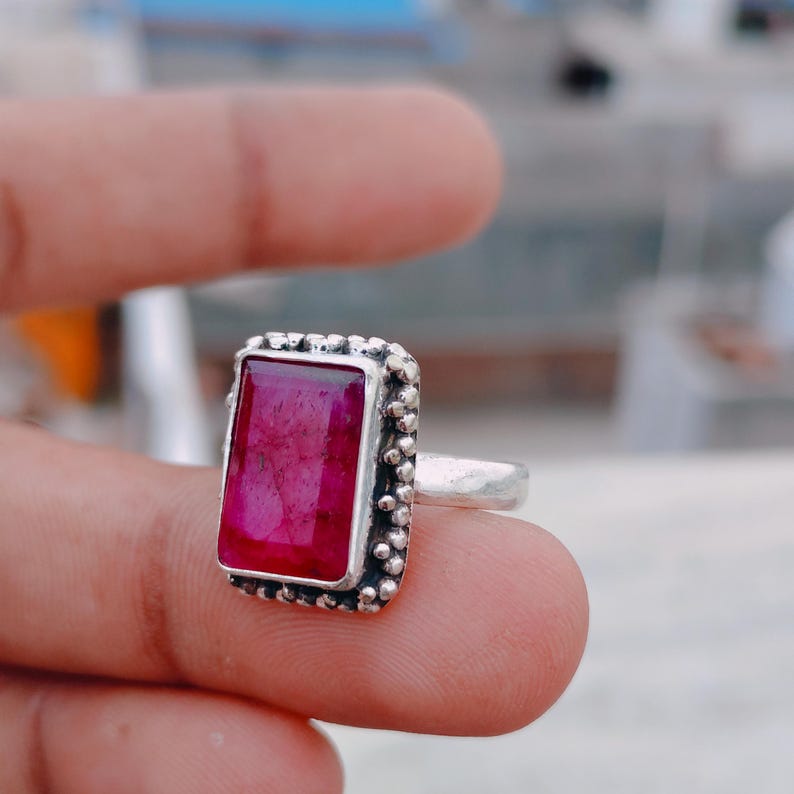 May include: A silver ring featuring a rectangular, deep pink gemstone set in a bezel with a beaded edge. The band is plain and metallic, showcasing a textured, handcrafted appearance around the stone setting, highlighting the vibrant color.
