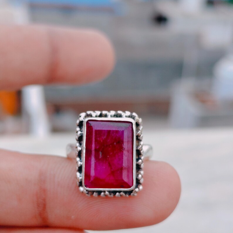 May include: A rectangular, deep pink gemstone ring set in a textured, oxidized silver band. The stone features a faceted surface with visible internal inclusions, framed by a decorative, beaded metal border that surrounds the entire setting.