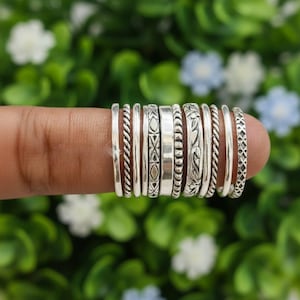 May include: A collection of ten silver rings arranged in a row, featuring various textures and patterns including twisted rope, smooth bands, geometric shapes, beaded details, and floral engravings, all displayed together on a single finger.