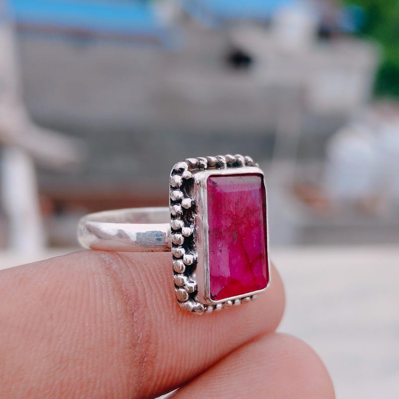 May include: A silver ring featuring a rectangular, faceted, deep pink gemstone set in a bezel. The metal setting surrounding the stone has a decorative, beaded texture along the edges, and the band is a smooth, polished silver metal.