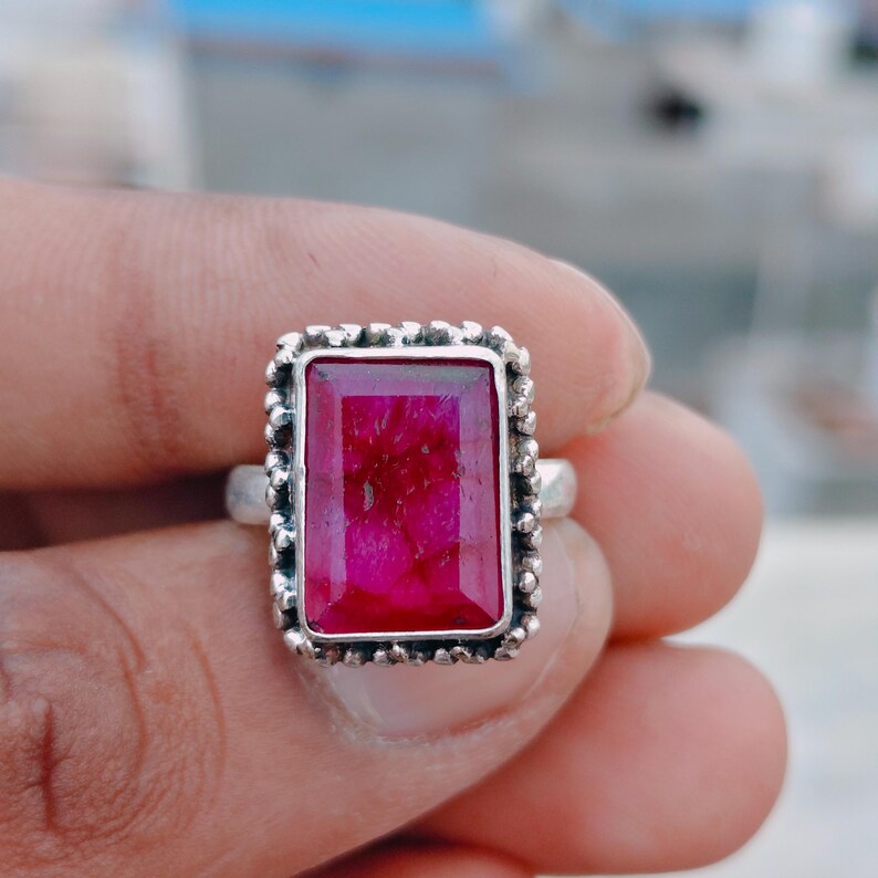 May include: A silver ring featuring a rectangular, faceted, deep pink gemstone set in a decorative, beaded metal frame. The stone has a vibrant, saturated color with visible internal inclusions, and the band is made of polished silver metal.