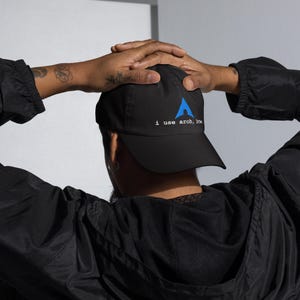 May include: Black baseball cap with a curved brim. The cap features the text "i use arch, btw." in white and a blue stylized upward-pointing arrow logo. The cap is worn by a person with tattoos on their arm.