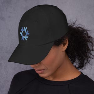 Könnte beinhalten: Eine schwarze Baseballkappe mit einem gestickten hexagonalen Logo auf der Vorderseite. Das Logo besteht aus sechs miteinander verbundenen hell- und mittelblauen Segmenten, die in einem kreisförmigen Muster angeordnet sind und ein geometrisches Schneeflocken-Design bilden.