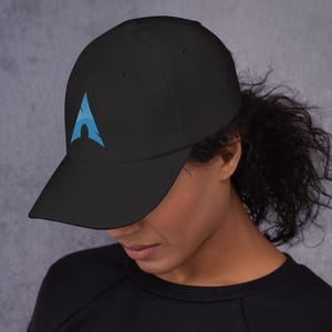 May include: Black baseball cap with a curved brim. The cap features a light blue logo on the front. The logo is an abstract design. The cap is made of a soft material.