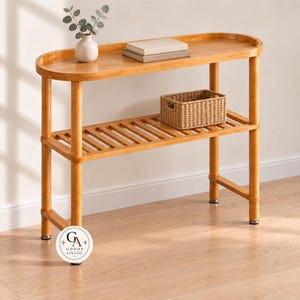 May include: A light brown wooden console table with an oval top and a slatted lower shelf. On the top surface sits a small white vase with green leaves and two stacked books. A woven rectangular basket rests on the lower shelf. A circular logo reads GA GOODS ALQASMI.