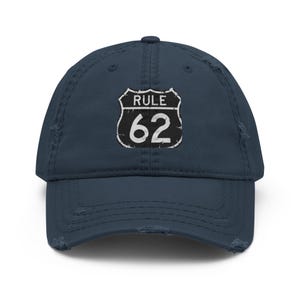May include: A navy blue distressed baseball cap featuring a white embroidered shield-shaped patch on the front. The patch displays the text RULE 62 in a bold, white font. The cap has a curved brim and frayed edges along the visor and crown.
