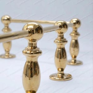 May include: A gold-colored, decorative towel rack with a polished finish. The rack features four vertical supports with a bulbous design, connected by horizontal bars. The base is circular, and the overall design is ornate.