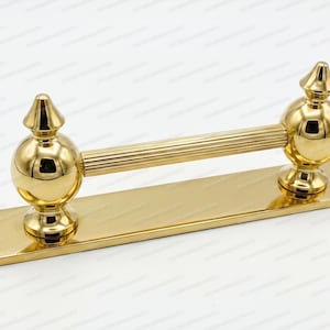 May include: A polished gold-tone metal door handle featuring a long, horizontal, ribbed cylindrical grip. The handle is mounted on a flat, rectangular base plate and includes two spherical supports topped with pointed finials at each end.
