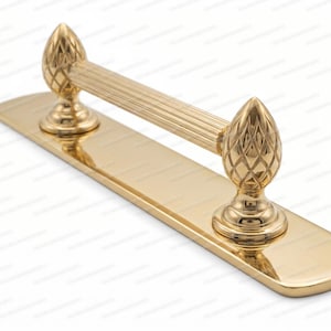 May include: A polished gold-tone metal door handle featuring a long, horizontally ribbed cylindrical grip. The handle is mounted on a rectangular backplate with rounded ends and is flanked by two decorative pineapple-shaped finials.