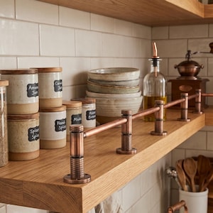 May include: A copper metal railing attached to the edge of a wooden kitchen shelf. The railing features cylindrical supports with a textured, cross-hatched pattern at the base and top, connected by a horizontal rod, designed to act as a guard rail.