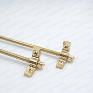 May include: Two brass-colored curtain rod brackets. Each bracket has a rectangular base with decorative detailing and a small, round finial. The rods are straight and extend horizontally.