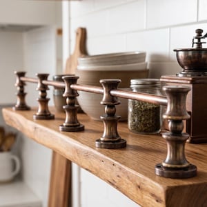 May include: A horizontal metal rail with a dark bronze finish, supported by five matching decorative vertical posts. The rail is mounted along the front edge of a wooden shelf, featuring a smooth, cylindrical rod and ornate, tiered supports.