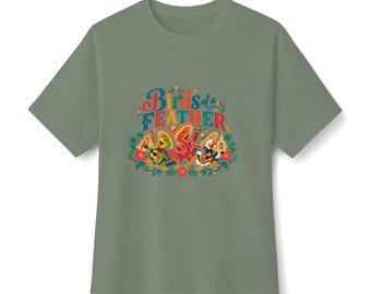 May include: A sage green short-sleeve t-shirt featuring a colorful graphic print with the text "Birds of a FEATHER". The design depicts three cartoon birds wearing sombreros and playing musical instruments, surrounded by flowers and foliage.