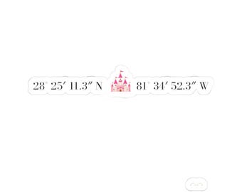 May include: A white die-cut sticker featuring the black text 28° 25' 11.3" N and 81° 34' 52.3" W, separated by a small, centered illustration of a pink castle with multiple turrets and flags, set against a plain white background.