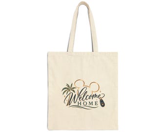 May include: A beige canvas tote bag featuring a graphic print with the text "Welcome HOME" in a dark script font. The design includes a gold-colored mouse ear silhouette, a green palm tree, wavy lines, and a small dark luggage tag icon.