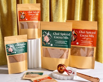 Puede incluir: Se muestran varias bolsas de Chai-Spiced Cocoa Mix, con etiquetas verdes, rojas y naranjas. También hay una cuchara de color cobre y pequeños paquetes de muestra. El fondo presenta una cortina dorada.