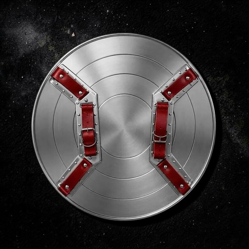 May include: A circular silver-toned metal shield featuring a series of concentric rings. The reverse side is equipped with two parallel metal brackets secured by rivets, each fitted with adjustable red leather straps and silver-toned metal buckles.
