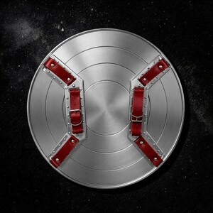 May include: A circular silver-toned metal shield featuring a series of concentric rings. The reverse side is equipped with two parallel metal brackets secured by rivets, each fitted with adjustable red leather straps and silver-toned metal buckles.