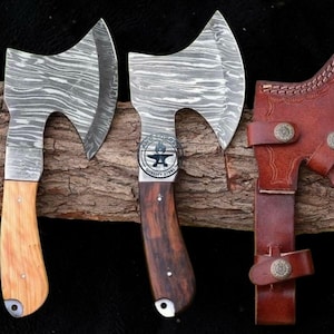 May include: Two hand axes with Damascus steel blades featuring wavy patterns and wooden handles. One handle is light-colored wood, the other is dark wood with a circular logo reading AHE FORGED QUALITY STEEL. A brown leather sheath is included.