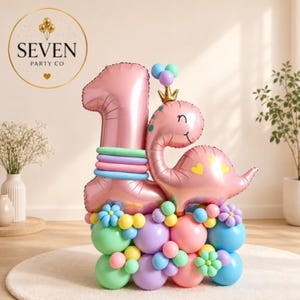 Op de afbeelding: Een roze folieballon in de vorm van het cijfer één, gecombineerd met een roze dinosaurusballon met een gouden kroon. De basis is opgebouwd uit een cluster van pastelkleurige latexballonnen in de tinten blauw, roze, geel en paars.