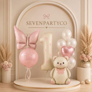 May include: A party balloon display featuring a pink butterfly balloon atop a round pink balloon, a large cream-coloured number one balloon, and a cream-coloured teddy bear balloon with a pink heart balloon. The text SEVENPARTYCO is visible.