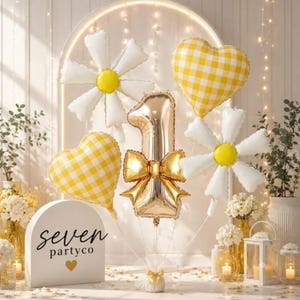 May include: A balloon bouquet featuring a large gold number one balloon with a gold bow, two white daisy-shaped balloons with yellow centers, and two yellow and white gingham heart-shaped balloons. A white arch sign reads seven partyco.