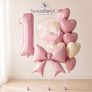 May include: A balloon arrangement featuring a large pink number one foil balloon, a clear bubble balloon with small pink bows inside, a large pink bow-shaped foil balloon, and a cluster of heart-shaped balloons in shades of pink and cream.
