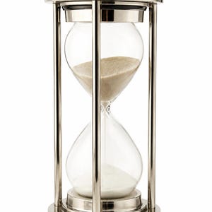 Puede incluir: Un reloj de arena de metal plateado con un bulbo de vidrio transparente lleno de arena beige fina. El marco consta de cuatro pilares verticales que conectan una base y una parte superior circulares, rematadas con esferas decorativas.