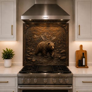 May include: A stainless steel gas range with a matching vent hood features a decorative bronze-toned metal backsplash panel. The panel displays a high-relief sculpture of a bear walking through a forest with mountains in the background.