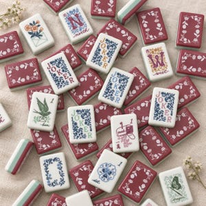 May include: A collection of rectangular mahjong tiles featuring white faces with colorful floral borders and illustrations. Some tiles display the word JOKER, directional letters like N for north and W for west, or images of a bird, envelope, and butterfly.