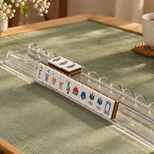 May include: A clear acrylic Mahjong tile rack holding a row of white tiles with colorful symbols, numbers, and the word JOKER. The rack features a series of rounded, semi-circular cutouts along the top edge, designed to support and display the tiles.