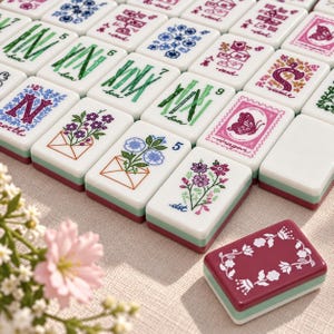 May include: A collection of rectangular mahjong tiles with white faces and green and maroon bases. The tiles feature colorful illustrations of bamboo stalks, flowers in envelopes, butterflies, and letters. One maroon tile with a white floral border sits separately in the foreground.