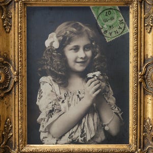 May include: A vintage-style portrait of a young person with curly hair and a flower, framed in an ornate gold-colored frame. A green postage stamp with the text MANA 14-19 07 is affixed to the upper right corner of the photograph.