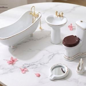 Puede incluir: Un juego de baño en miniatura de cerámica blanca que incluye una bañera con grifos dorados, un lavabo de pedestal, un inodoro con asiento marrón, un pequeño espejo redondo y un portavelas. El texto intothedollhouse es visible en la esquina.