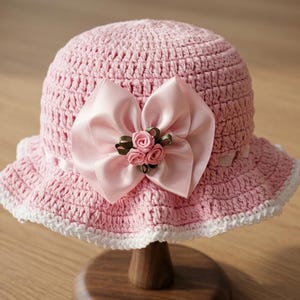 May include: A light pink crocheted bucket hat featuring a wide satin ribbon bow at the front. The center of the bow is decorated with three small fabric roses in shades of pink and green. The hat has a white crocheted trim along the brim edge.