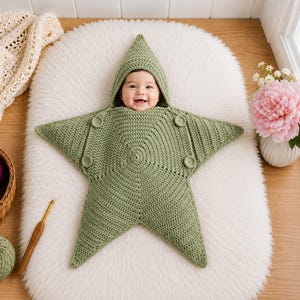 May include: A sage green crocheted baby swaddle blanket shaped like a five-pointed star with an integrated hood. The garment features a textured stitch pattern and four circular buttons positioned on the front to secure the star-shaped flaps.