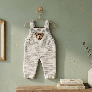 May include: A cream and grey speckled crochet overall featuring a brown bear face appliqué on the chest. The garment has two shoulder straps with grey buttons at the front and ribbed cuffs at the leg openings, hanging on a gold metal hanger.
