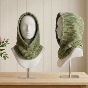 May include: A sage green crochet hood with a textured ribbed pattern that covers the head and neck. The hood is displayed on two white mannequin heads, showing both a front-facing view and a side profile view of the knitted headwear accessory.