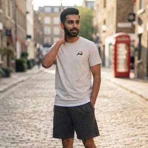 May include: A light beige short-sleeved crew neck t-shirt featuring a small embroidered graphic of a three-wheeled vehicle on the left chest, paired with dark charcoal grey shorts that reach above the knee with a simple hemline.