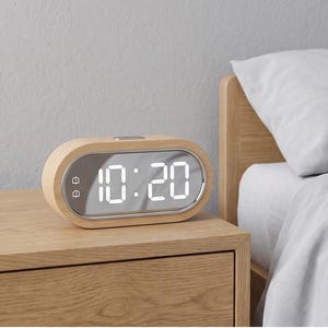 Peut inclure: Un réveil numérique avec une finition en bois clair. L'horloge affiche l'heure 10h20 en chiffres blancs. L'horloge a une face miroir et est posée sur une table de chevet en bois.