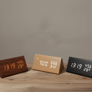 May include: Three wooden digital alarm clocks in triangular shapes. One is dark brown, one is light brown, and one is black. Each clock displays the time, humidity, and temperature in Celsius. The clocks are on a wooden surface.