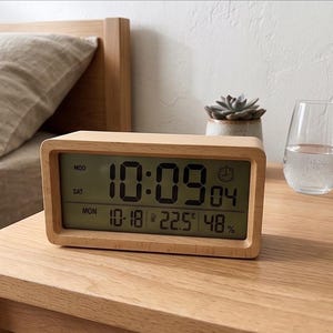 Pode incluir: Um despertador digital de madeira mostrando a hora 10:09, com data, temperatura e humidade. O relógio está numa mesa de cabeceira de madeira, ao lado de um copo de água e de uma pequena suculenta.