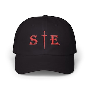 May include: A black baseball cap featuring an embroidered red design on the front. The design consists of the letter S, a vertical sword icon in the centre, and the letter E, forming the text STE in a bold, serif font with red stitching.