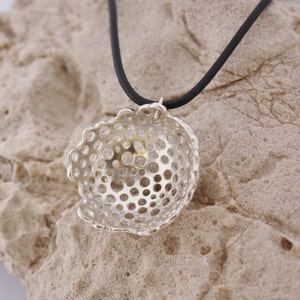 May include: A silver pendant necklace with a delicate, openwork design. The pendant is shaped like a small bowl with a circular pattern of holes. The necklace is on a black cord.
