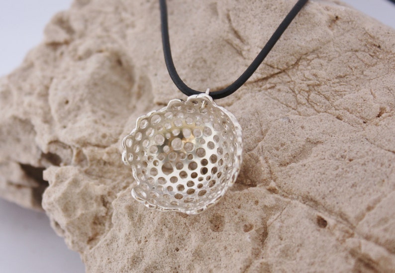 May include: A silver pendant necklace with a delicate, openwork design. The pendant is shaped like a small bowl with a circular pattern of holes. The necklace is on a black cord.