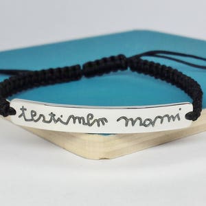 May include: Black braided bracelet with a silver bar charm engraved with the words "terribile mammi".