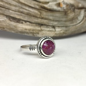 Peut inclure: Une bague en argent avec une pierre précieuse rose foncé sertie dans une bélière texturée. La bague a une bande simple avec un motif texturé.